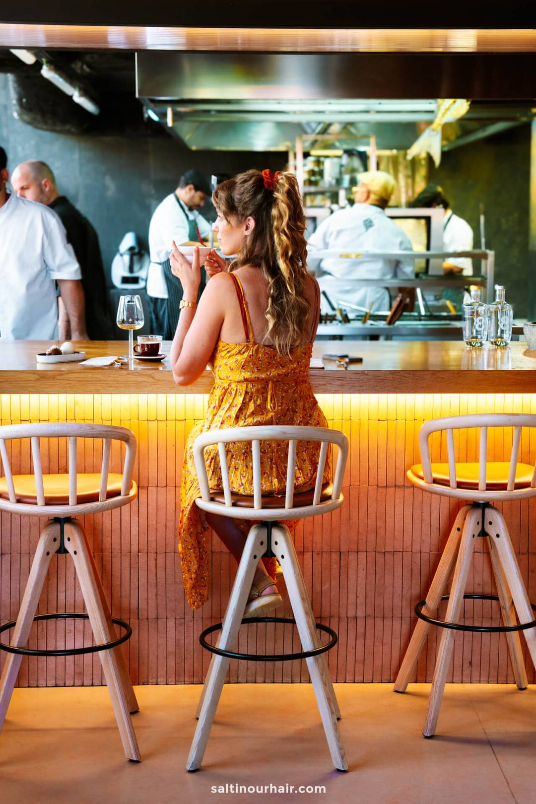 A woman in a yellow dress sits at a bar counter, holding a drink, with chefs working in the background&mdash;a stylish scene capturing the lively dining atmosphere among the top things to do in Funchal, madeira