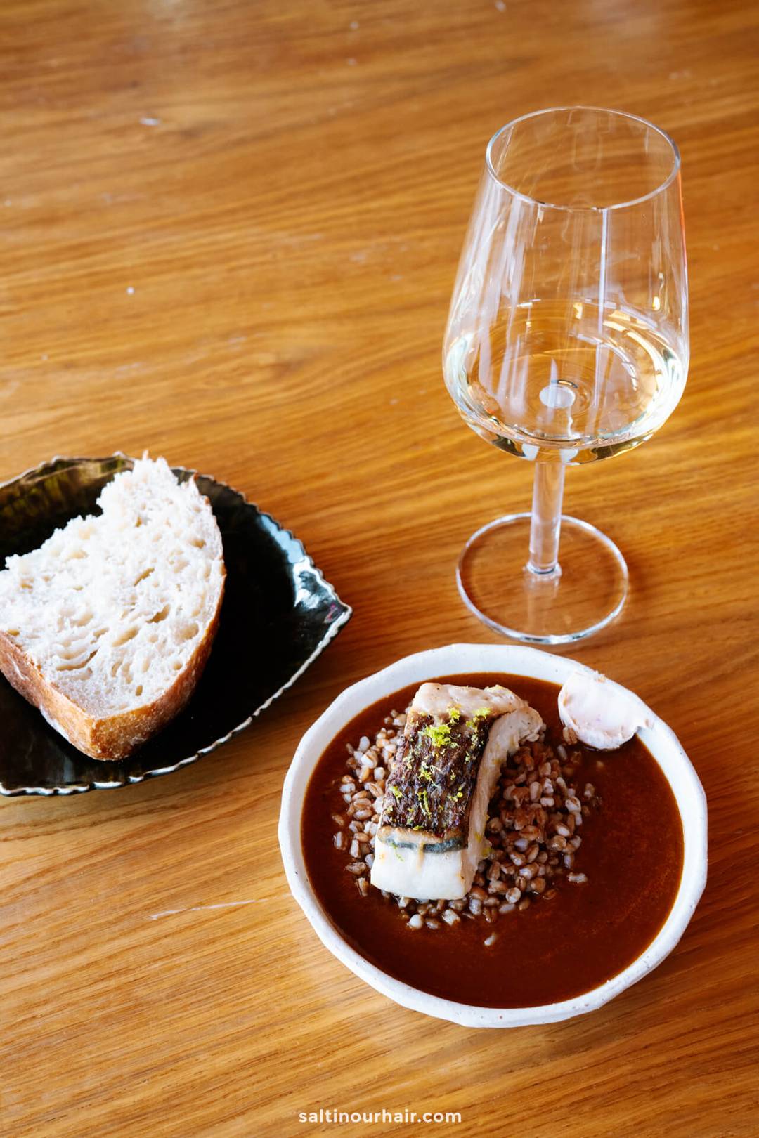 A plate with fish on grains in sauce, a slice of bread on a side dish, and a glass of white wine on a wooden table&mdash;a perfect way to enjoy local flavors while exploring things to do in Funchal, madeira