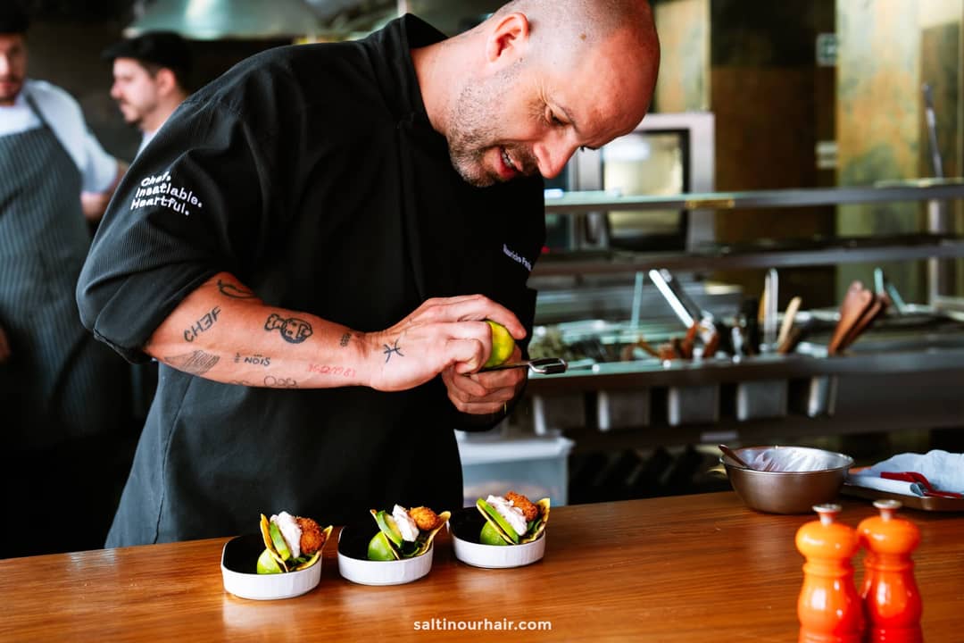 A chef garnishes small dishes of food in a professional kitchen in Funchal, with kitchen utensils and staff visible in the background&mdash;perfect for anyone exploring things to do in Funchal, madeira