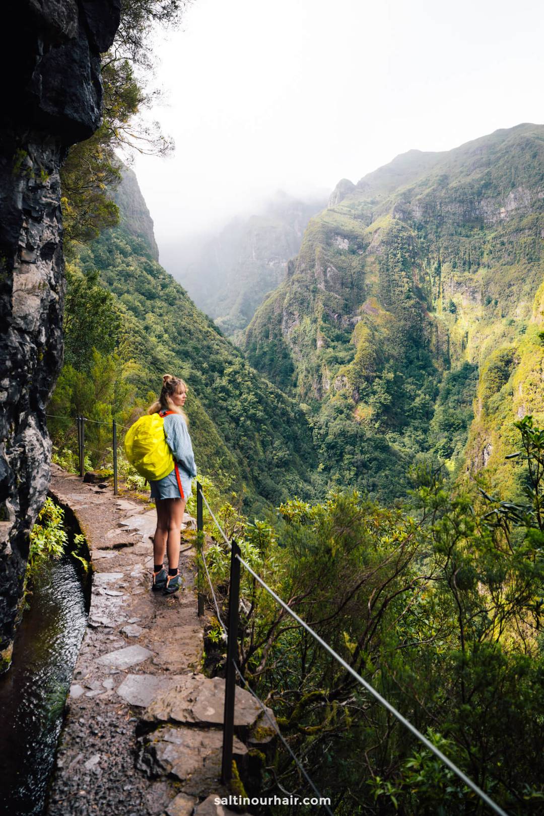Een persoon met een gele rugzak staat op een smal bergpad naast een weelderige groene vallei en rotsachtige kliffen, terwijl hij de schilderachtige route verkent zonder zich zorgen te maken over de wandelkosten op Madeira.