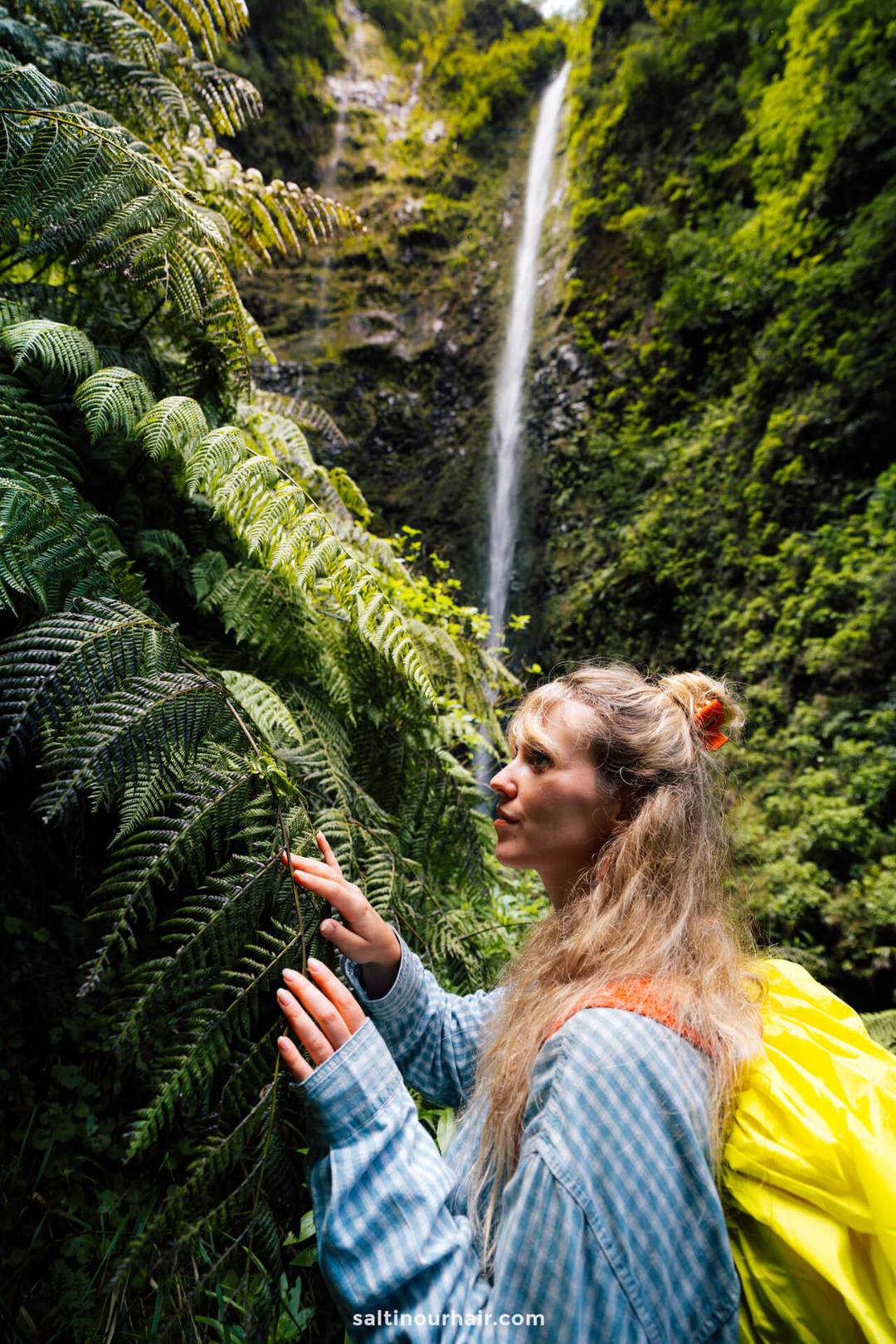 Een vrouw met een gele rugzak bekijkt varenbladeren in een weelderig, groen bos bij een hoge waterval, terwijl ze de schoonheid van de natuur verkent en rekening houdt met de wandelkosten op Madeira.
