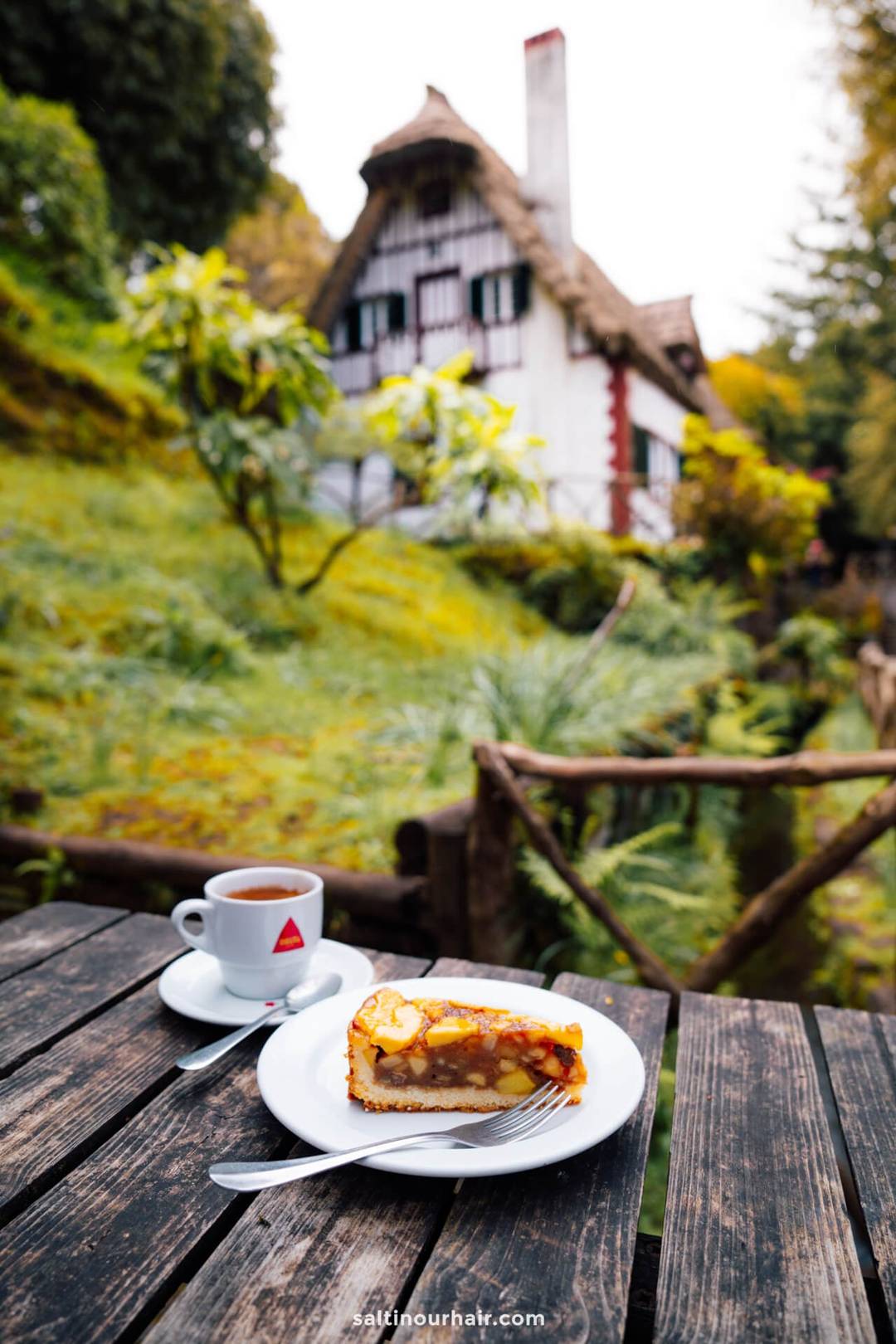 Een stuk appeltaart en een kop thee op een houten tafel buiten, met een rustiek huis en groene tuin op de achtergrond &mdash; perfect om de wandelkosten op Madeira te bespreken na een dag in de natuur.