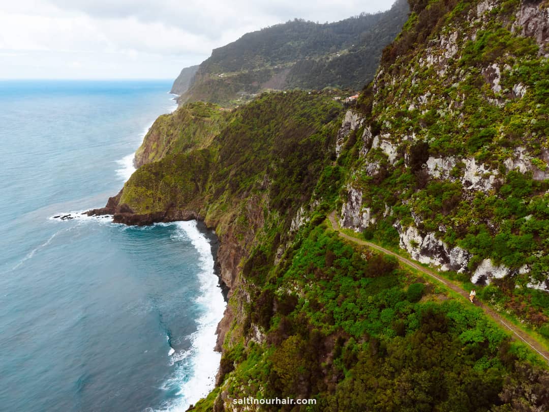 A narrow hiking trail runs along steep green cliffs overlooking the ocean, with waves crashing against the rocky shoreline below&mdash;one of the best hikes Madeira has to offer.