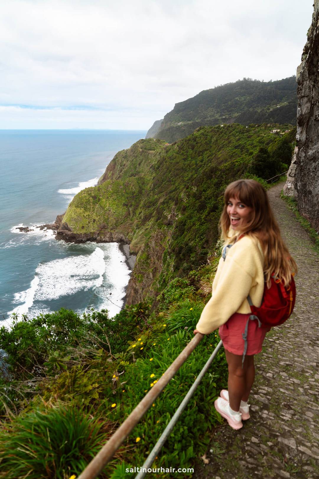 A woman with a red backpack stands on a stone path along a steep coastal cliff, overlooking the ocean and green hills under a cloudy sky&mdash;one of the best hikes Madeira has to offer.