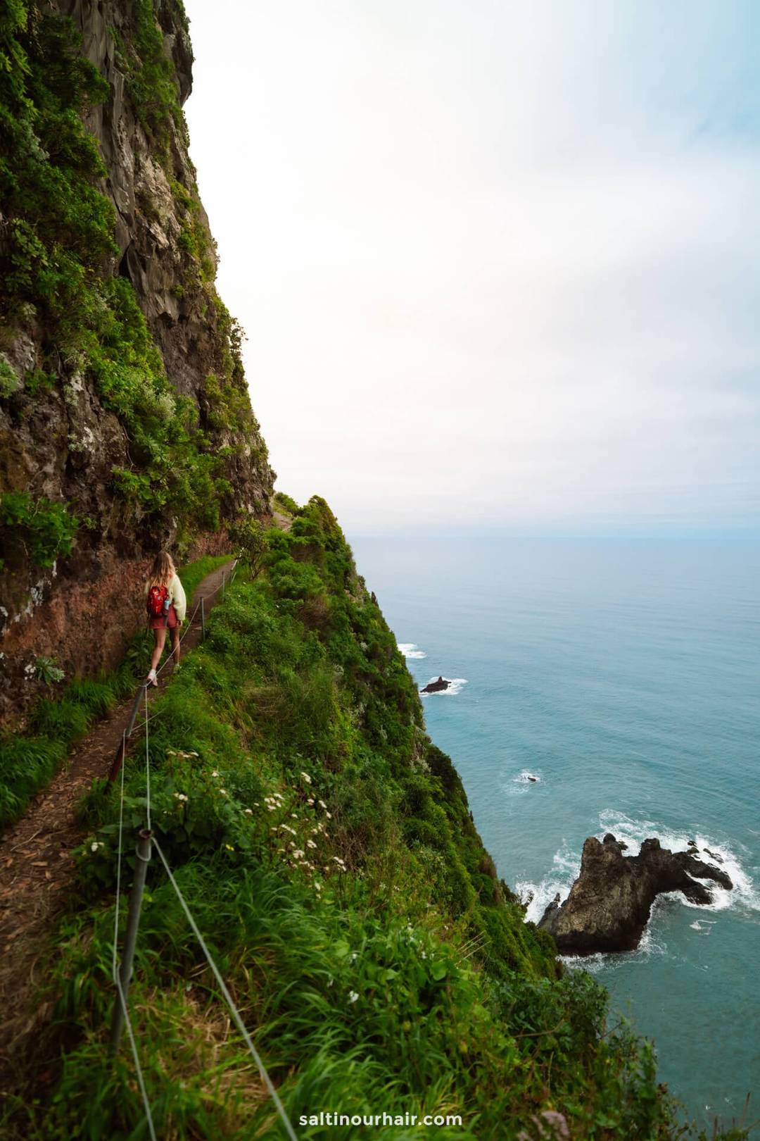 Person walking along a narrow trail on a steep, green cliff above the ocean&mdash;cloudy sky overhead and rocky outcrops below&mdash;captures the breathtaking scenery of one of the best hikes Madeira has to offer.