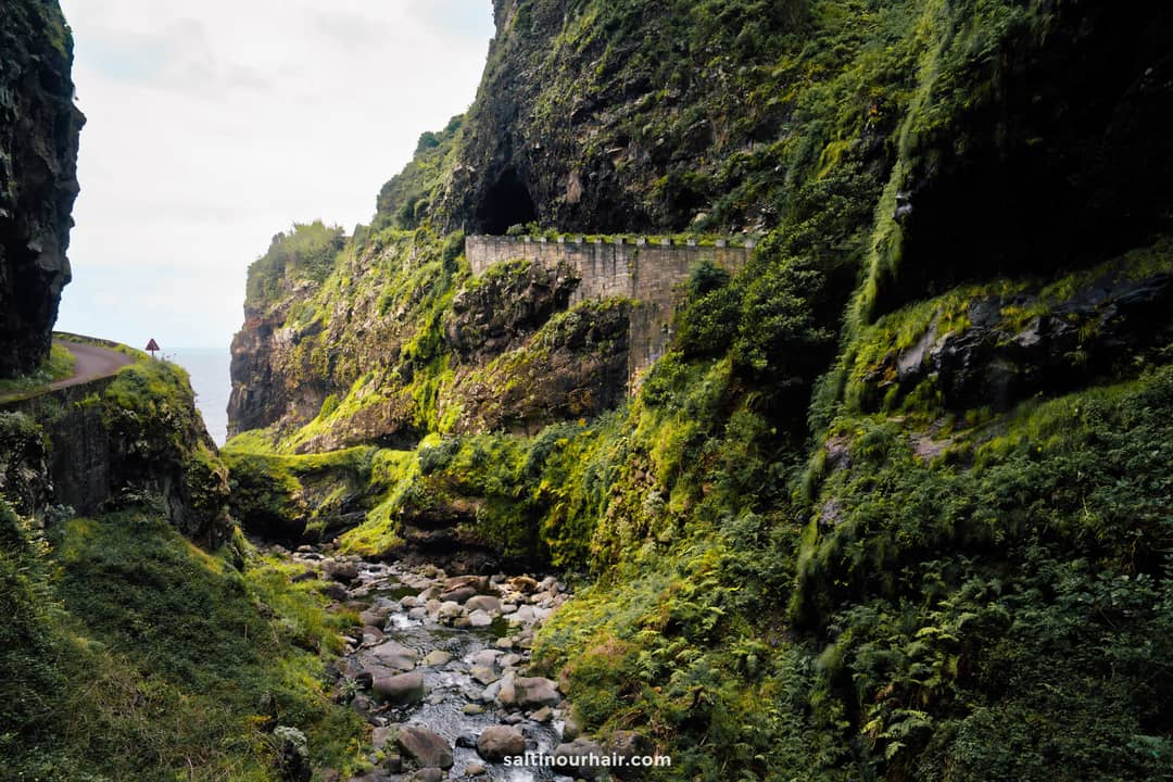 Narrow road and tunnel carved into steep, rocky green cliffs above a rocky stream, with a person walking in the distance &mdash; Ribeira do Inferno (ER101) &mdash; a glimpse of the best hikes Madeira has to offer.