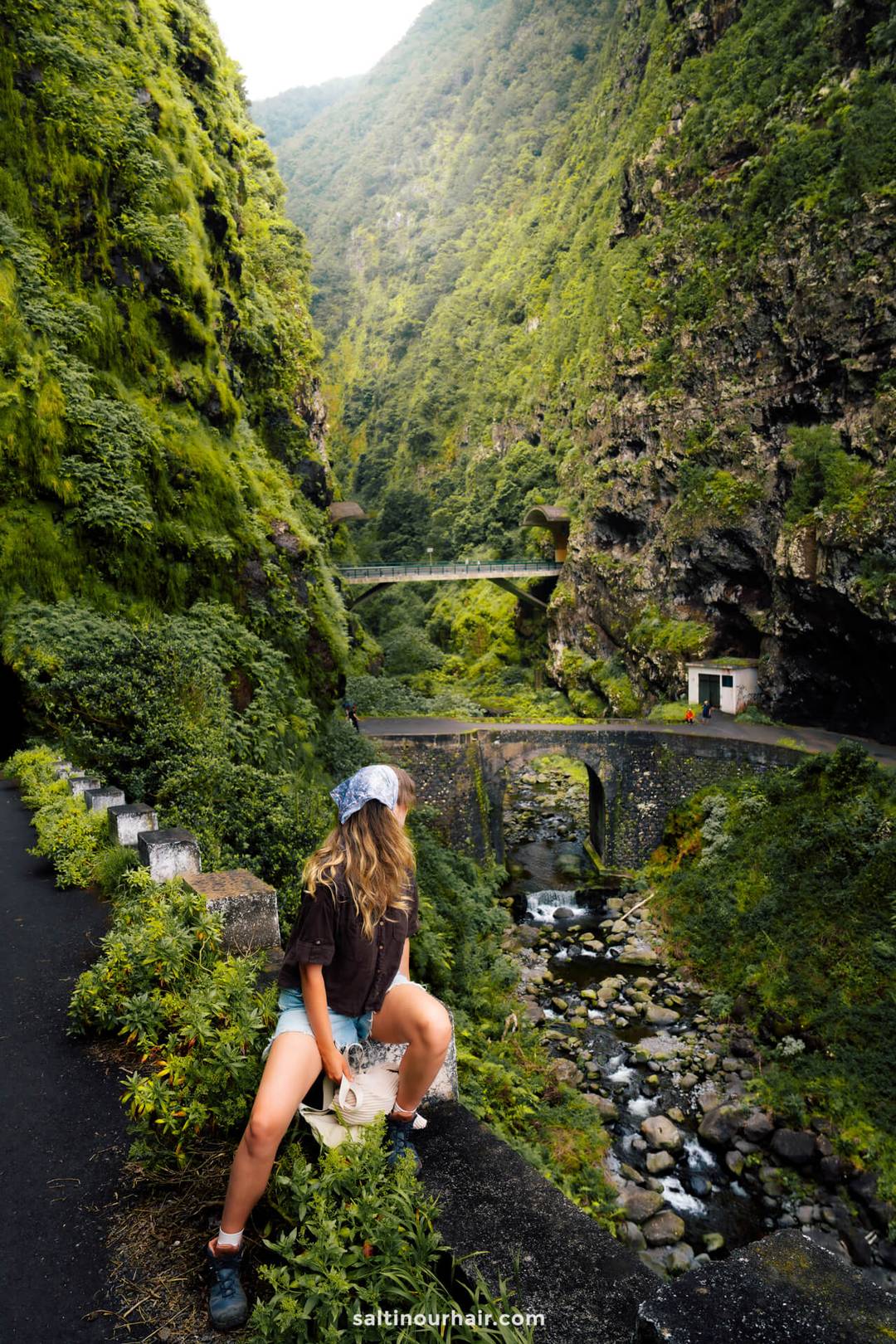 A person with long hair and a headscarf sits on a rock beside a winding mountain road called 