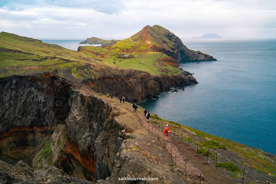 Hikers walk along a narrow, rocky coastal trail called Ponta do São Lourenço Hike &mdash; PR8 &mdash;one of the best hikes Madeira offers&mdash;with steep cliffs and green hills overlooking the ocean under a partly cloudy sky.