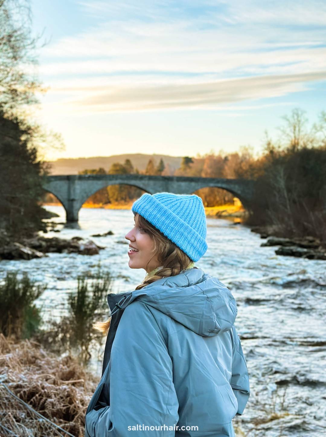 vrouw met blauwe muts die geniet van het Schotse platteland, rivier en brug