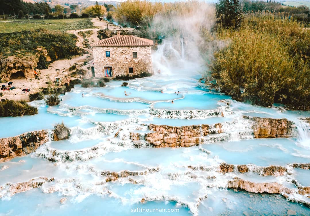 Aerial view of natural thermal hot springs with cascading turquoise pools and a stone building surrounded by greenery; a must-see stop for any Italy itinerary 2 weeks, where people are bathing in the pools.
