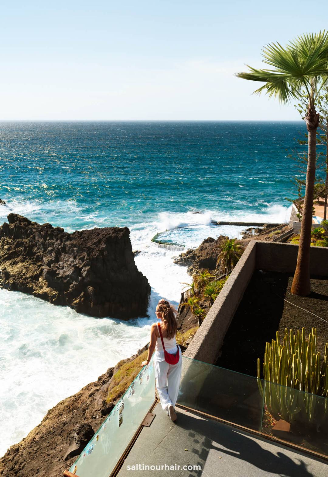 A woman in white stands on a balcony overlooking the rocky cliffs of Los Gigantes Tenerife, with palm trees and the oceans waves crashing below under a clear sky.