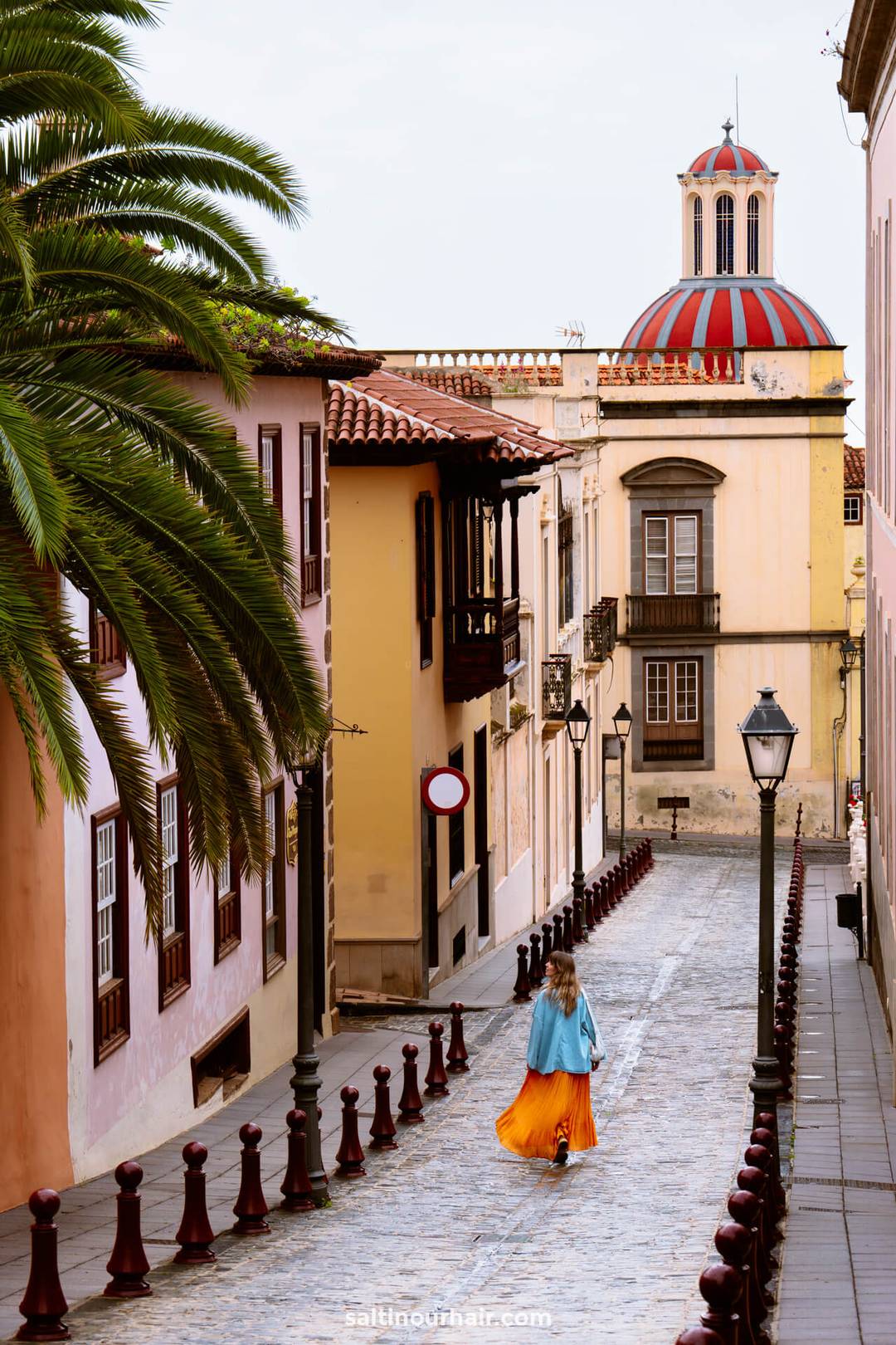 A woman in a blue jacket and orange skirt walks down a cobblestone street in La Orotava, lined with historic buildings and palm trees, with a domed building in the background.