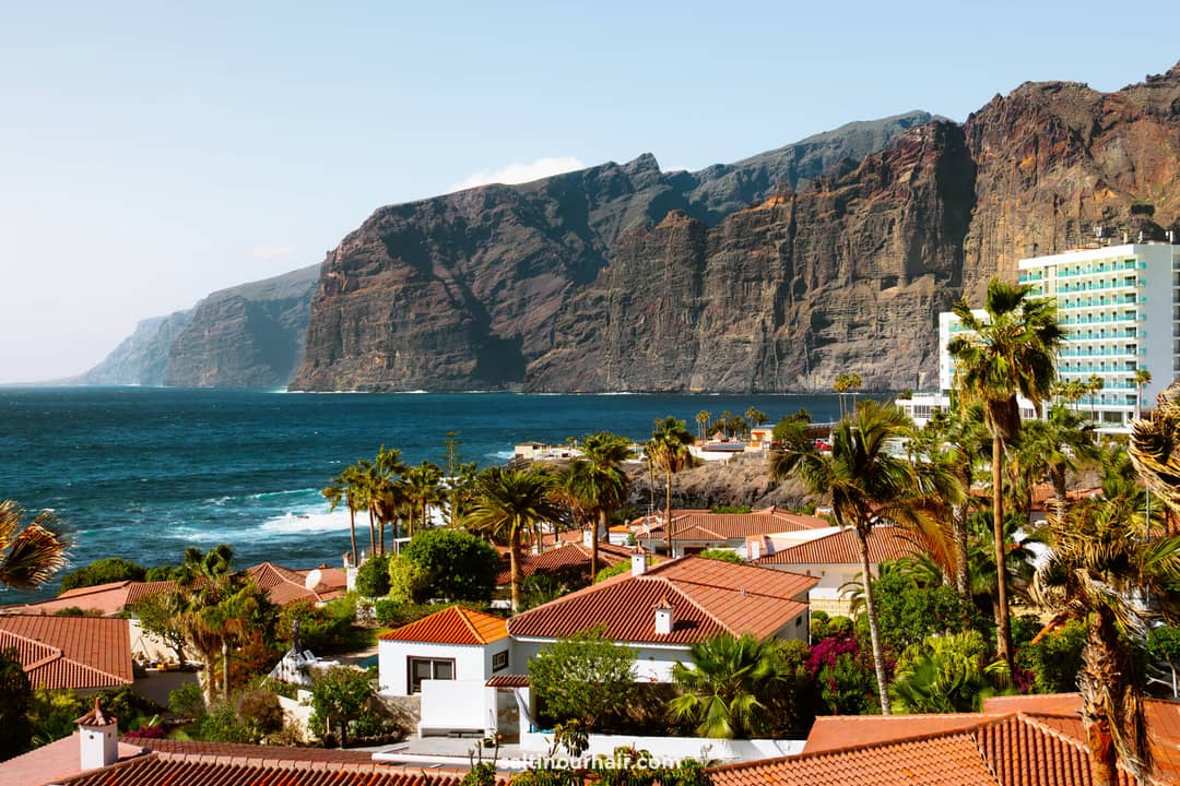 Coastal village with red-roofed houses and palm trees in the foreground, set against the dramatic cliffs of Los Gigantes Tenerife and the sparkling ocean in the background.