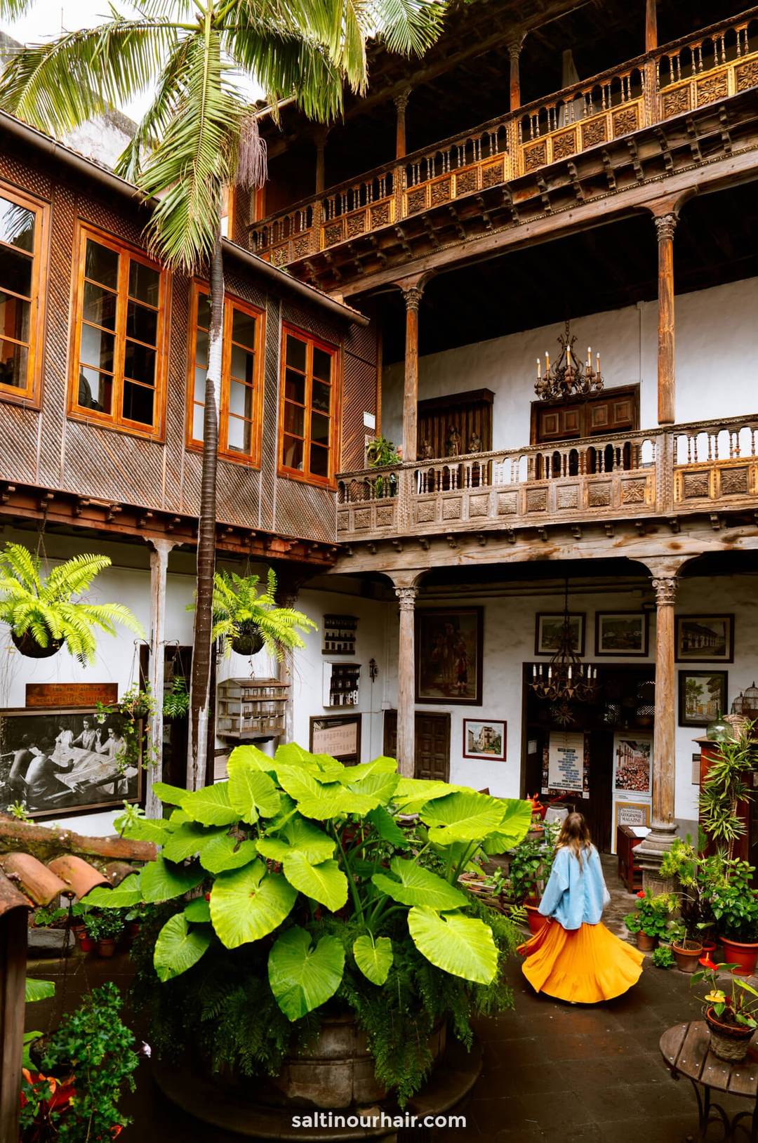 Woman in an orange dress stands in a lush courtyard at Casa de los Balcones La Orotava, surrounded by wooden balconies, potted plants, and historical decor in a colonial-style building.