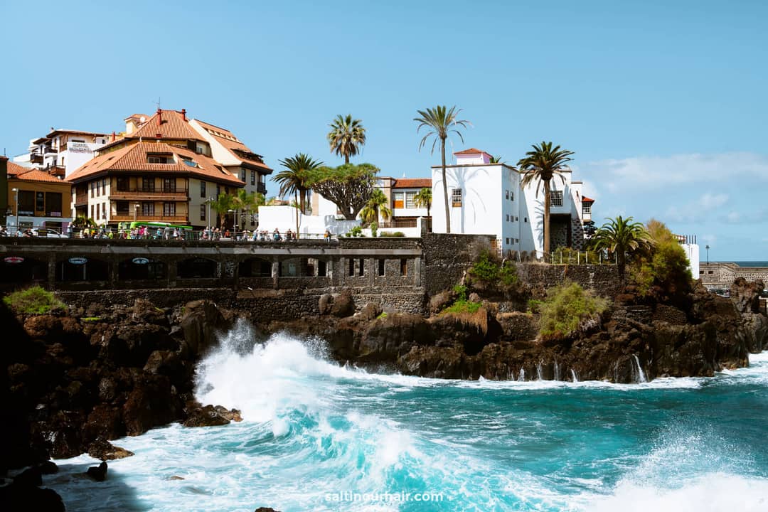 Waves crash against rocky cliffs below coastal buildings and palm trees under a clear blue sky along the scenic Paseo de San Telmo, Puerto de la Cruz, Tenerife.