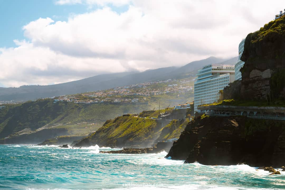 A seaside cliff with waves crashing below, modern buildings on the cliffside, and rolling hills in the background under a partly cloudy sky&mdash;much like the scenic views near Playa Jard&iacute;n, one of the top things to do in Puerto de la Cruz, Tenerife.
