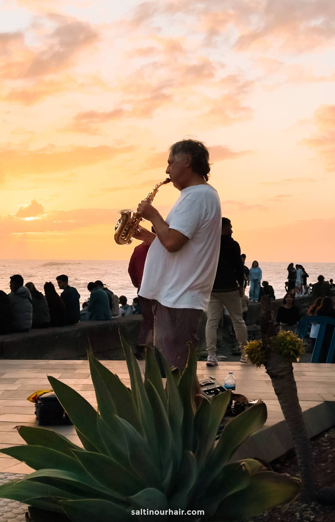 A man plays a saxophone outdoors at sunset near the ocean, with people sitting and standing in the background&mdash;an unforgettable moment to add to any Tenerife itinerary.