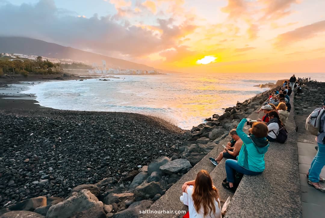 People sit on a stone seawall and watch the sunset over a rocky beach and ocean, with mountains and buildings in the background&mdash;the perfect spot to relax during your Tenerife itinerary.