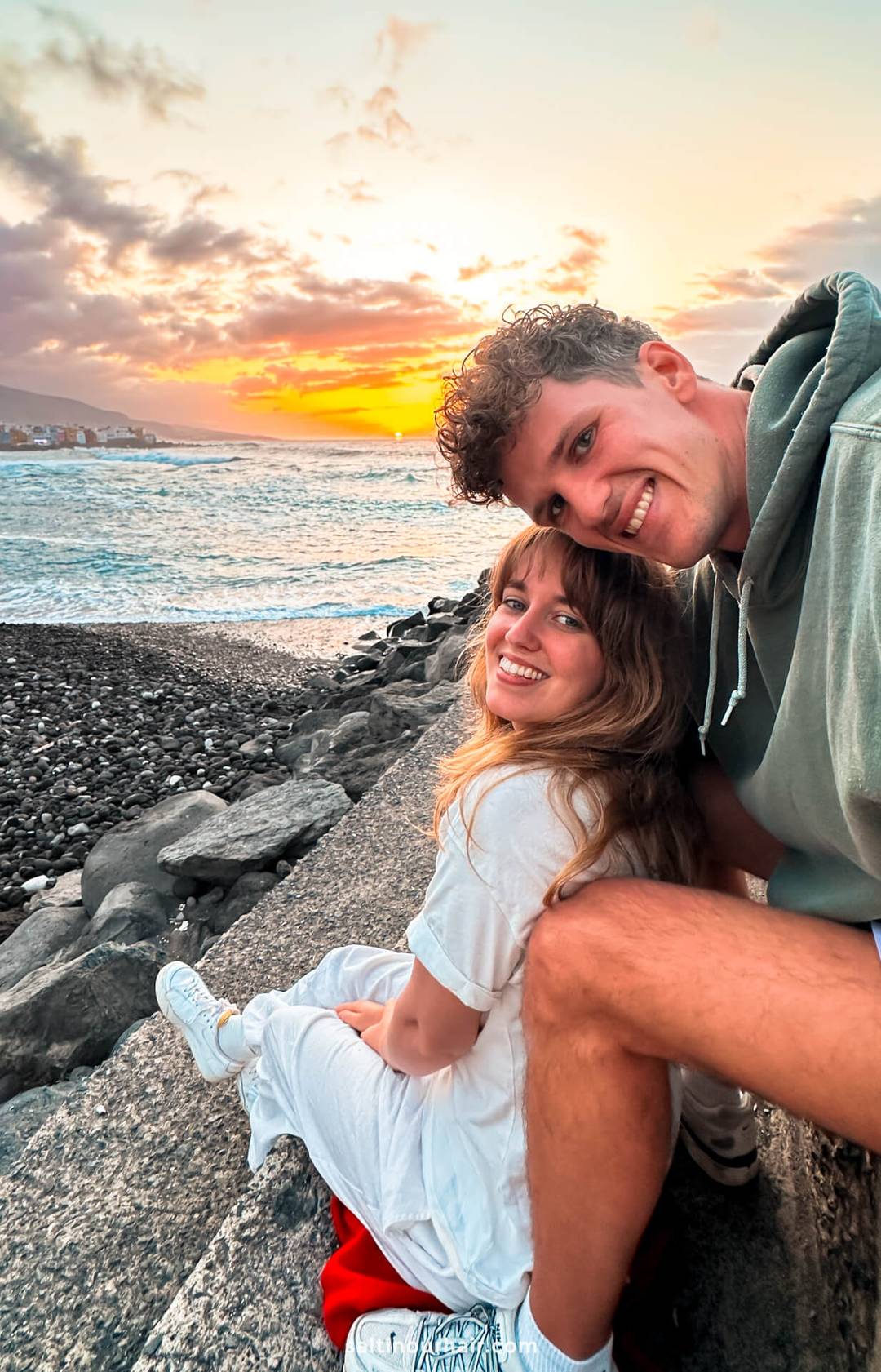 A smiling couple sits on a stone ledge by a rocky beach at sunset, the ocean and colorful clouds behind them&mdash;capturing a perfect moment for any Tenerife itinerary.