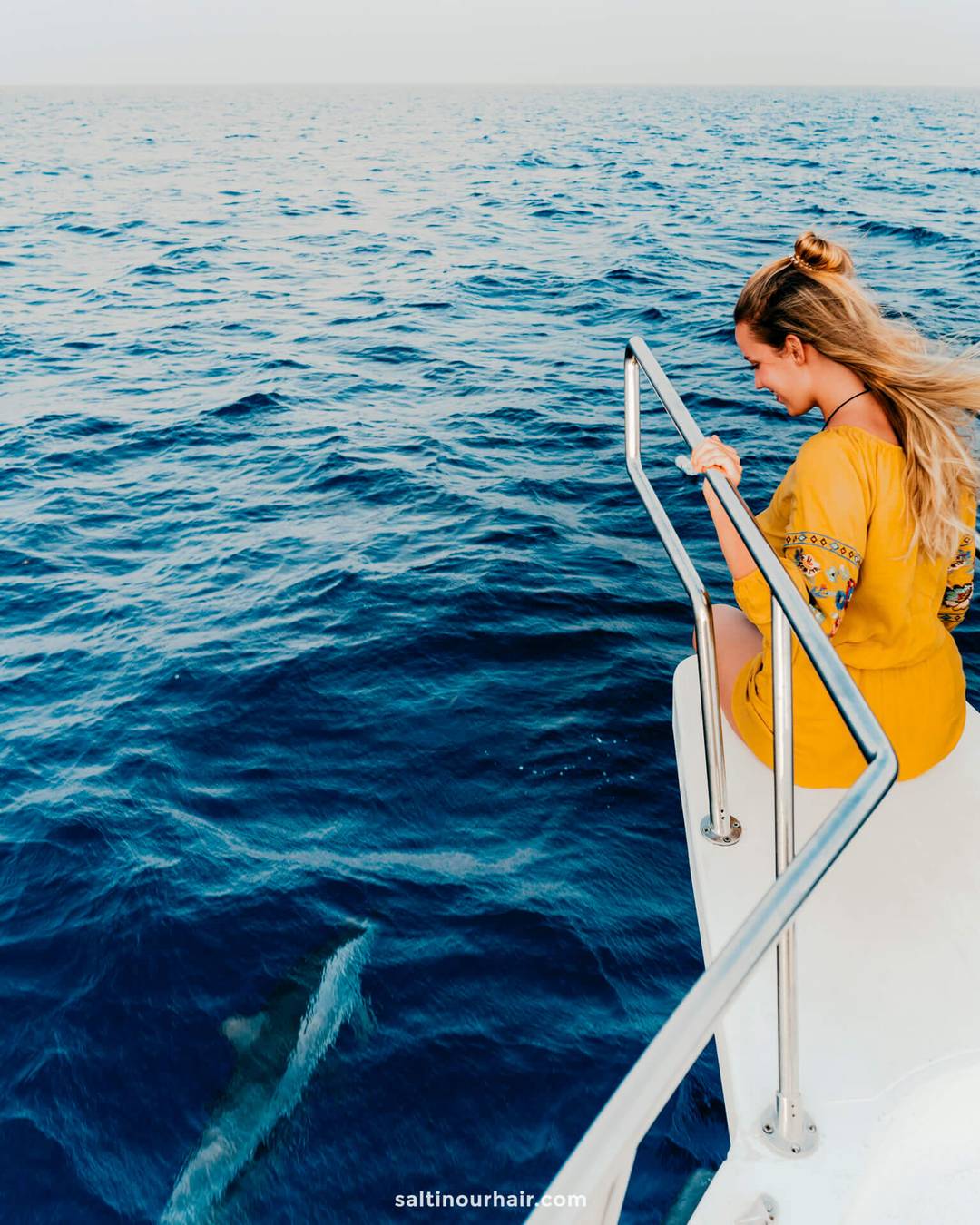 A woman in a yellow dress sits on the edge of a boat during a whale watching tour Los Gigantes Tenerife, looking down at a dolphin swimming in the blue ocean below.