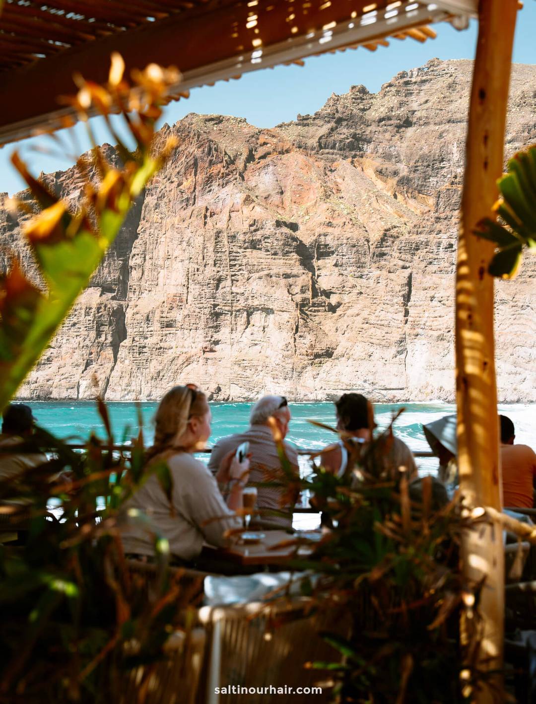 People seated at an outdoor restaurant at Los Gigantes Tenerife, enjoying drinks with views of turquoise water and towering rocky cliffs, all framed by lush plants and wooden beams.