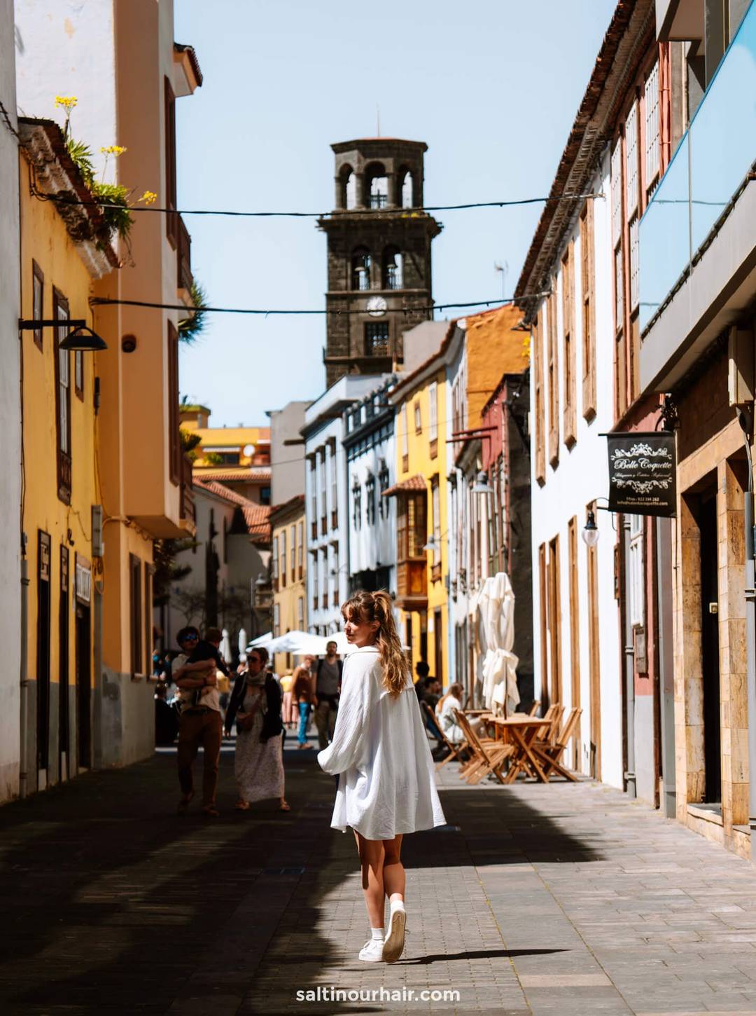 Een vrouw in een witte jurk loopt door een zonovergoten, smal straatje met kleurrijke gebouwen in San Crist&oacute;bal de La Laguna, Tenerife, met een stenen klokkentoren op de achtergrond.