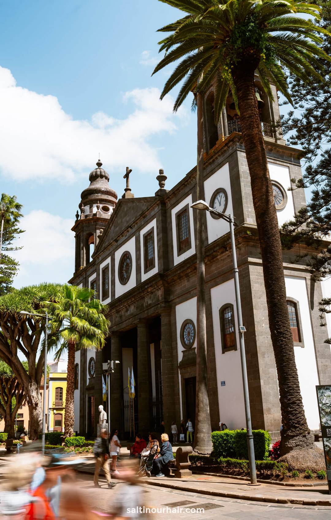 De kathedraal van La Laguna in San Crist&oacute;bal de La Laguna, Tenerife, met dubbele torens naast palmbomen, terwijl mensen voorbijlopen en op bankjes zitten. 