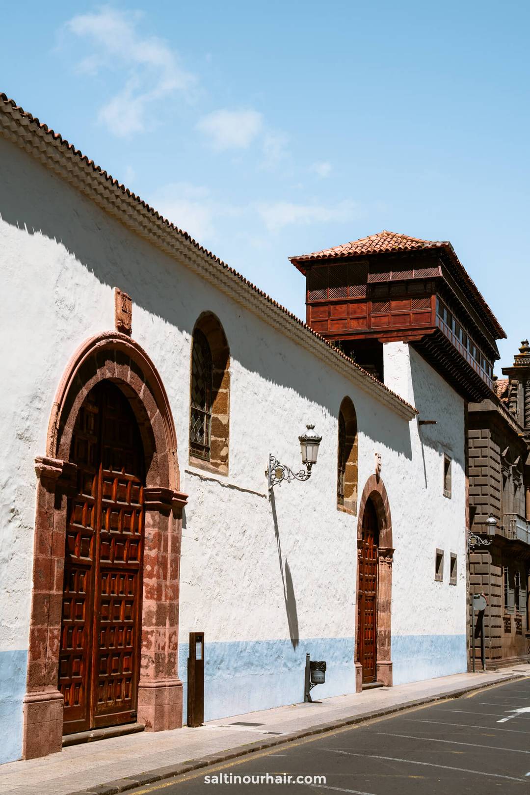 Een wit gestuukt gebouw met rode stenen bogen, houten deuren en een balkon, gelegen aan een rustige straat onder een blauwe lucht in San Crist&oacute;bal de La Laguna, Tenerife.