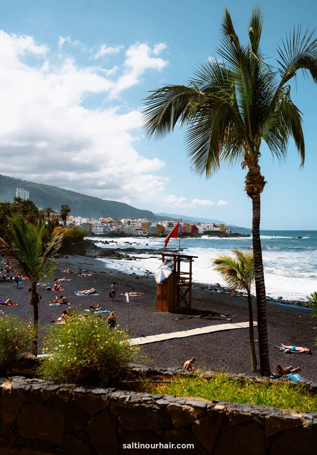 People sunbathe on Playa Jardin in Puerto de la Cruz, Tenerife, a black sand beach with palm trees, gentle waves, and colorful buildings in the distance under a partly cloudy sky.