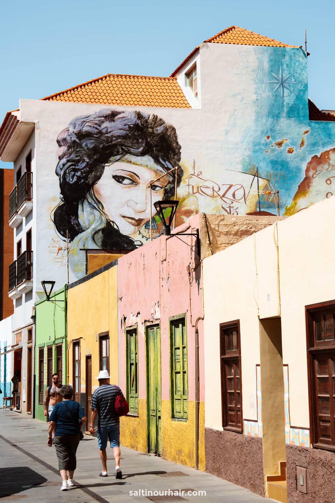 Street scene in La Ranilla Neighborhood, Puerto de la Cruz, Tenerife, with colorful buildings, two people walking, and a large mural of a womans face painted on the upper wall against a clear sky.