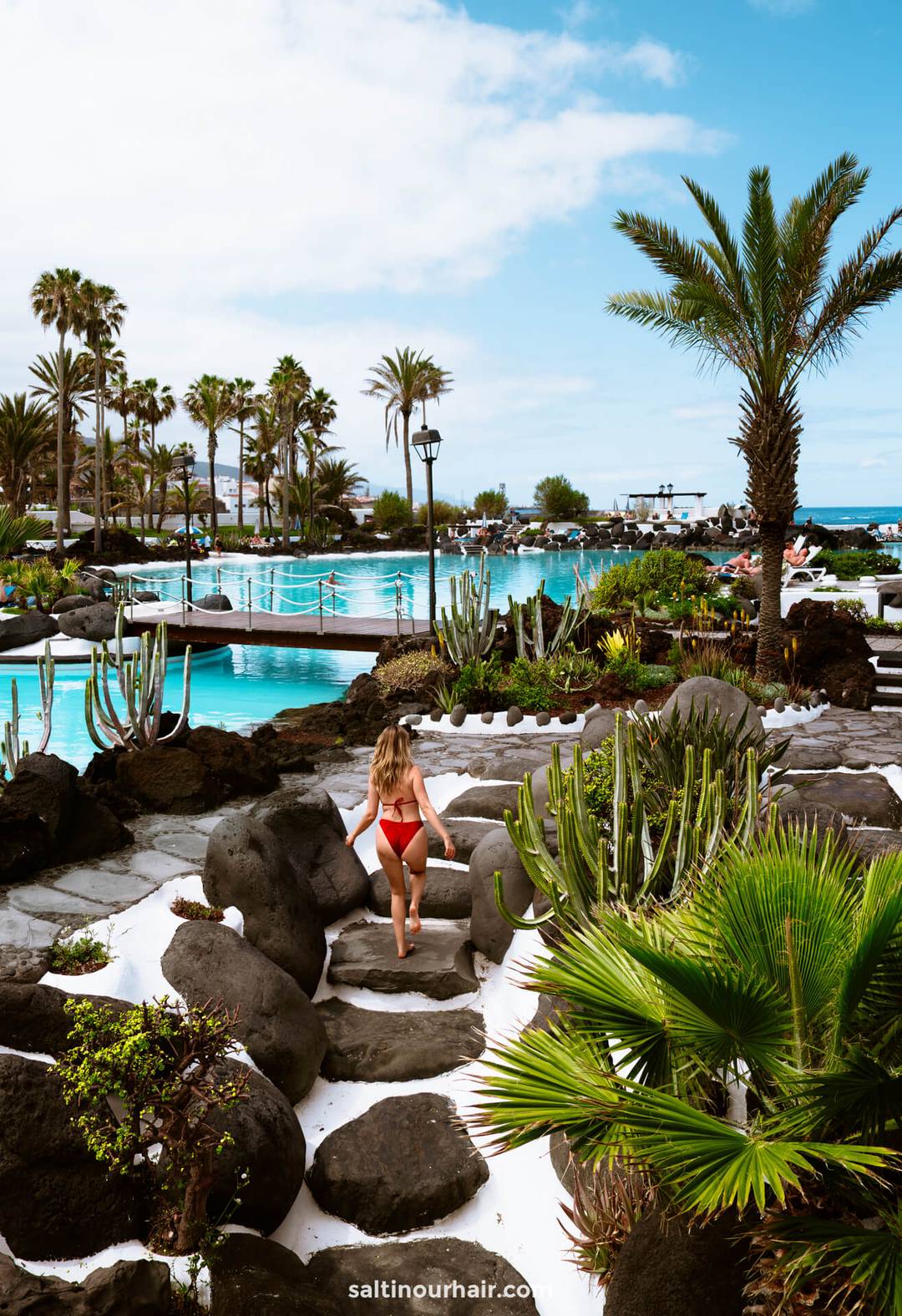A woman in a red bikini walks down stone steps toward a large, turquoise swimming pool at Lago Marti&aacute;nez in Puerto de la Cruz, Tenerife, surrounded by palm trees and tropical plants under a partly cloudy sky.