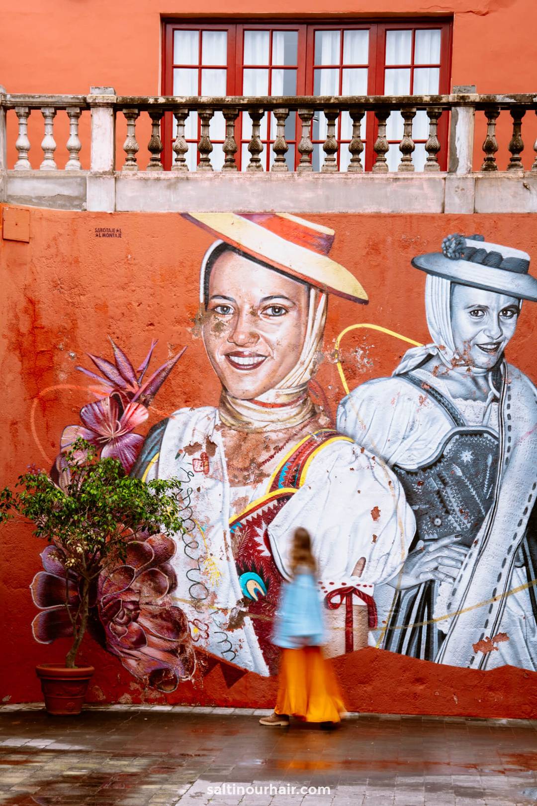 A mural depicting two women in traditional attire is painted on an orange wall; a blurred person in colorful clothing walks past a potted plant&mdash;capturing the vibrant street culture among the many things to do Tenerife offers.