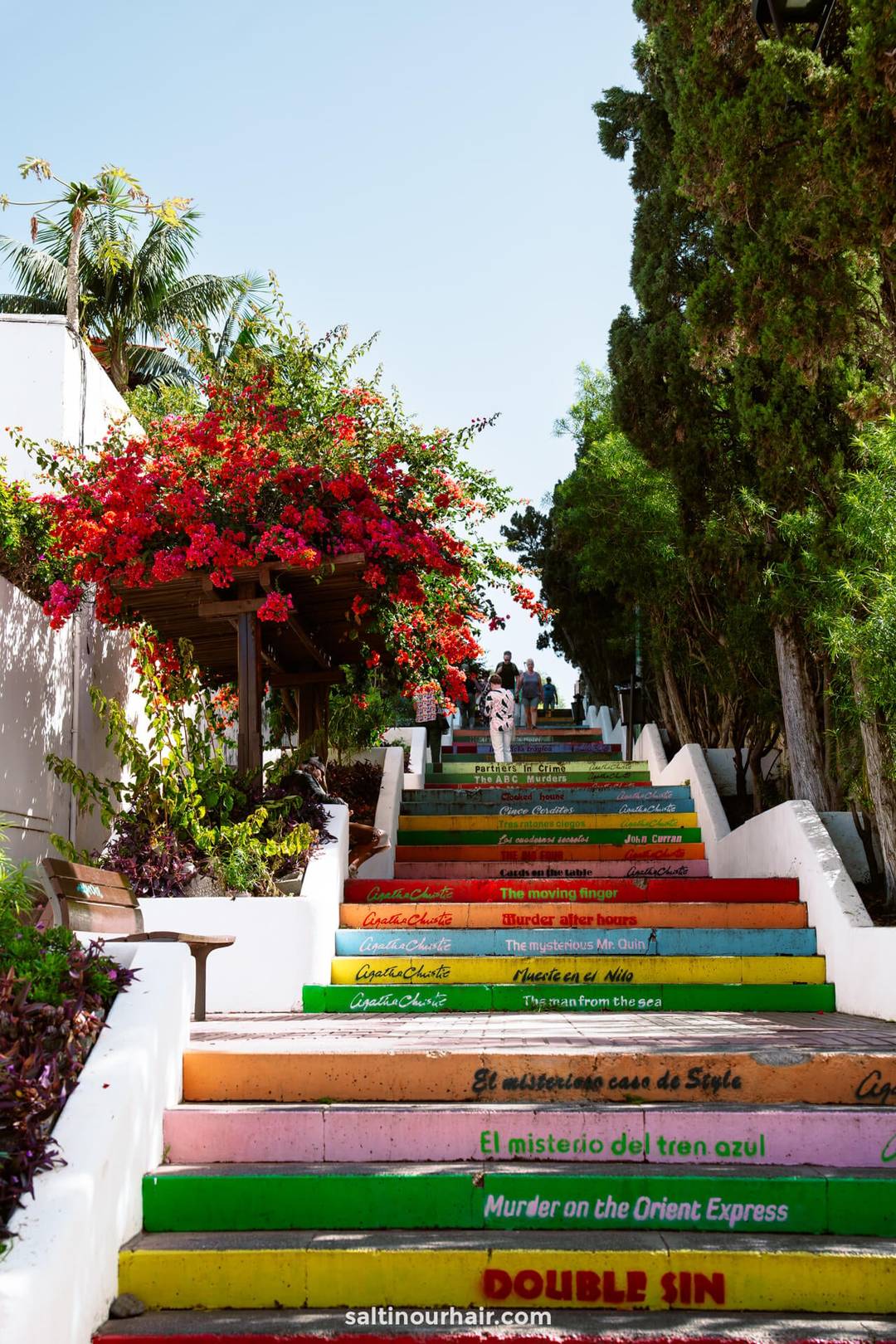 A colorful outdoor staircase in Puerto de la Cruz, Tenerife, known as the Agatha Christie Painted Stairs, features steps painted in vibrant hues and labeled with famous detective novels; trees and flowers line both sides.