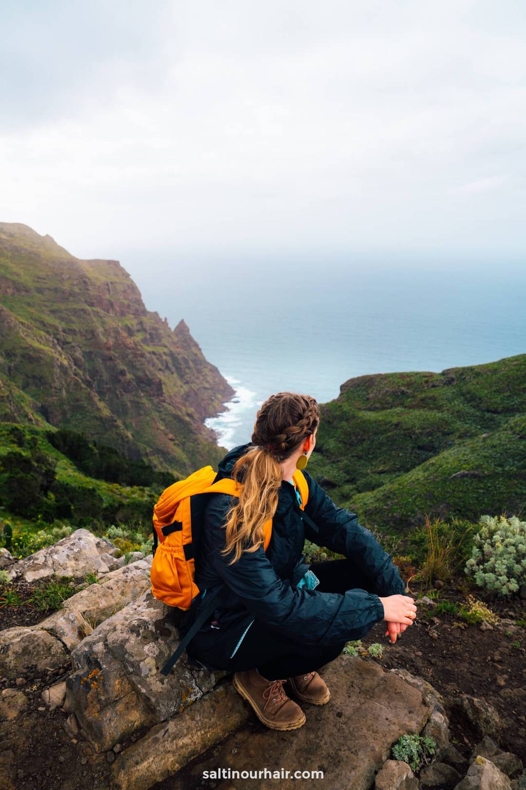Vrouw met een gele rugzak en gevlochten haar zit op een rots en kijkt uit over het groene berglandschap en de oceaan van Anaga Rural Park, de perfecte dagtrip vanuit San Crist&oacute;bal de La Laguna.