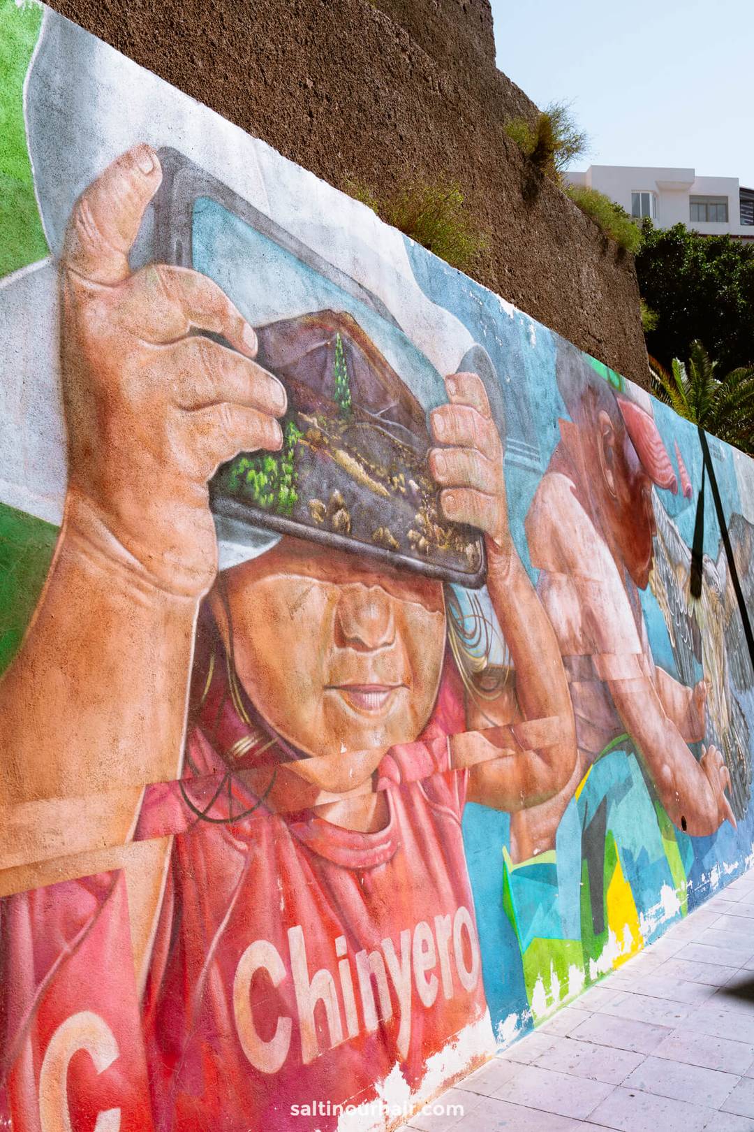A mural depicts a child holding a VR headset with a nature scene inside and the word Chinyero on their shirt, set against a textured outdoor wall in Los Gigantes, Tenerife.