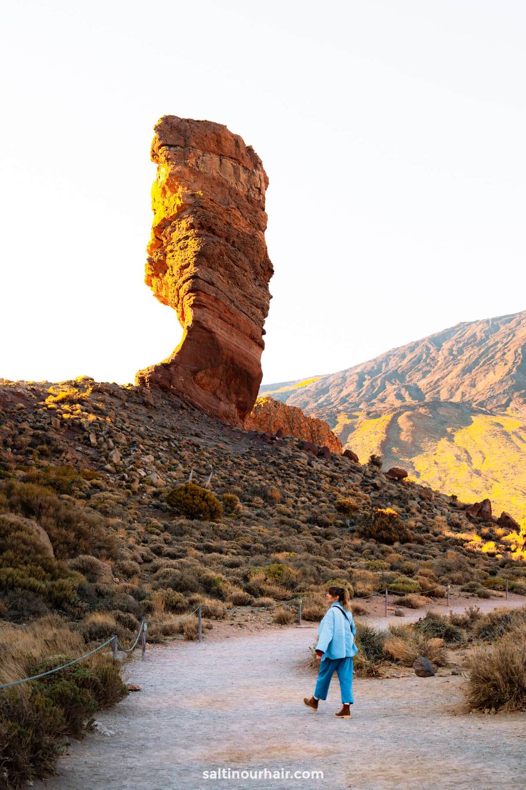 A women walks on a dirt path toward the tall, uniquely shaped Roques de Garc&iacute;a in Teide National Park, Tenerife, surrounded by dry, rocky terrain and distant hills.