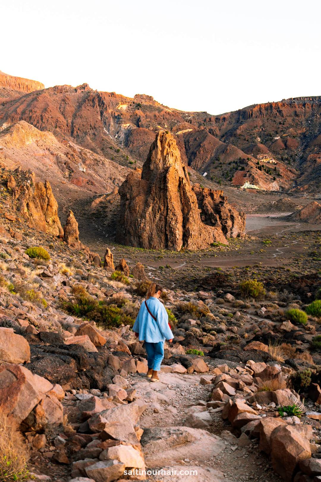 A women wearing a blue jacket walks on a rocky path toward a tall rock formation in the mountainous, arid landscape of Teide National Park, Tenerife.