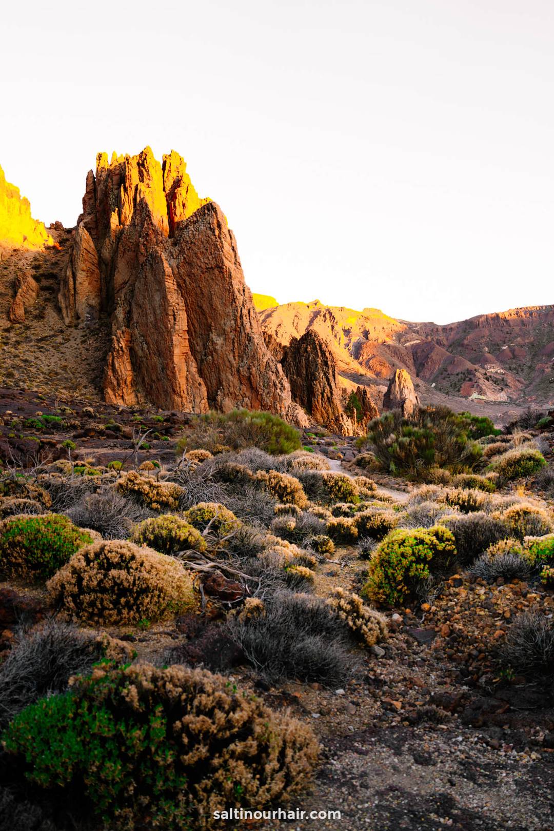 Rocky desert landscape with jagged cliffs illuminated by sunlight, dry shrubs, and sparse vegetation in the foreground, reminiscent of Teide National Park Tenerife.