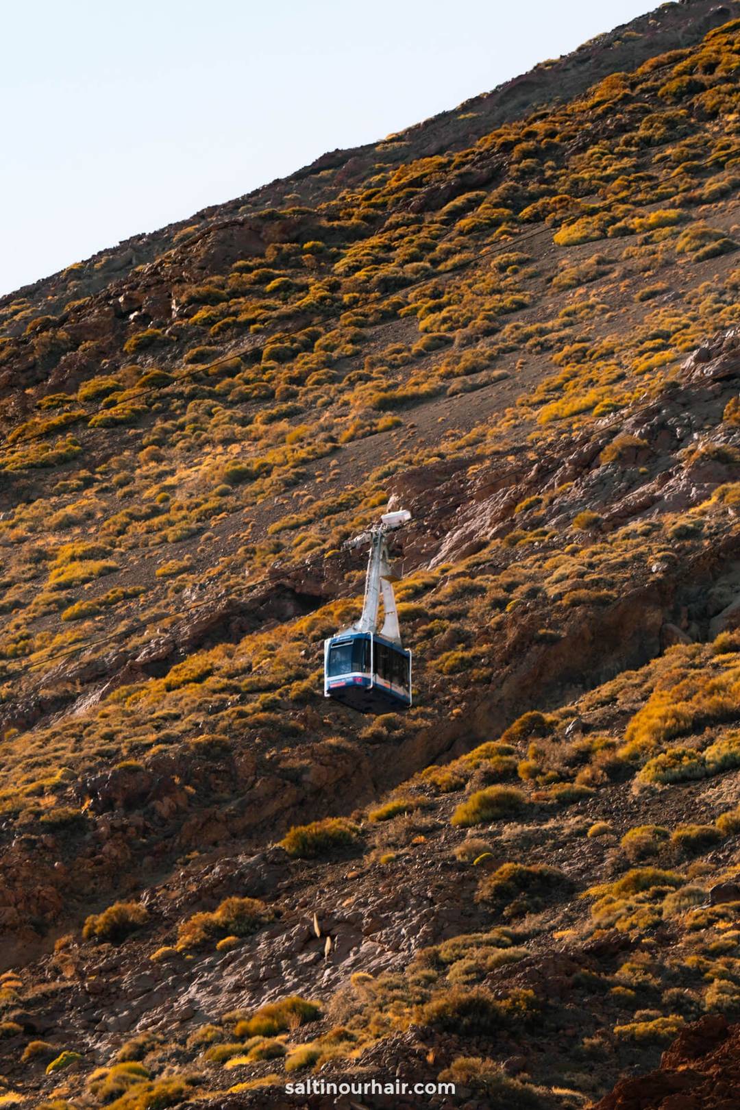 A cable car travels up a steep, rocky hillside covered with patches of dry grass and shrubs under bright sunlight in Teide National Park tenerife