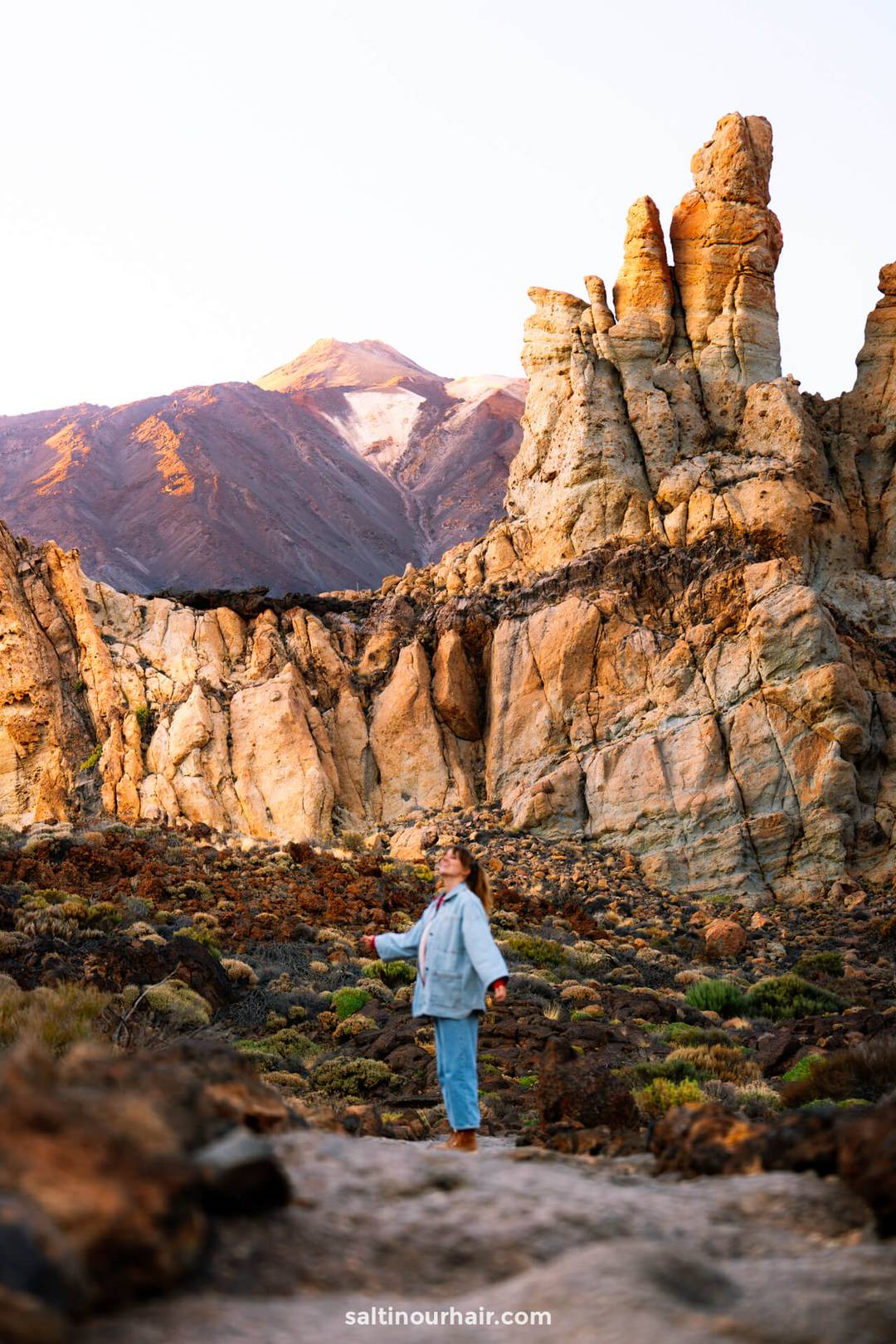 A woman stands on rocky terrain in Teide National Park, Tenerife, surrounded by rugged, sunlit rock formations with the majestic teide volcano in the background.