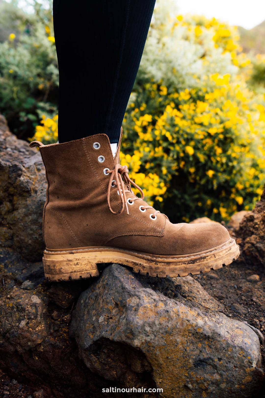 A person wearing brown lace-up hikinh boots stands on a rock, with yellow flowers and greenery in the background