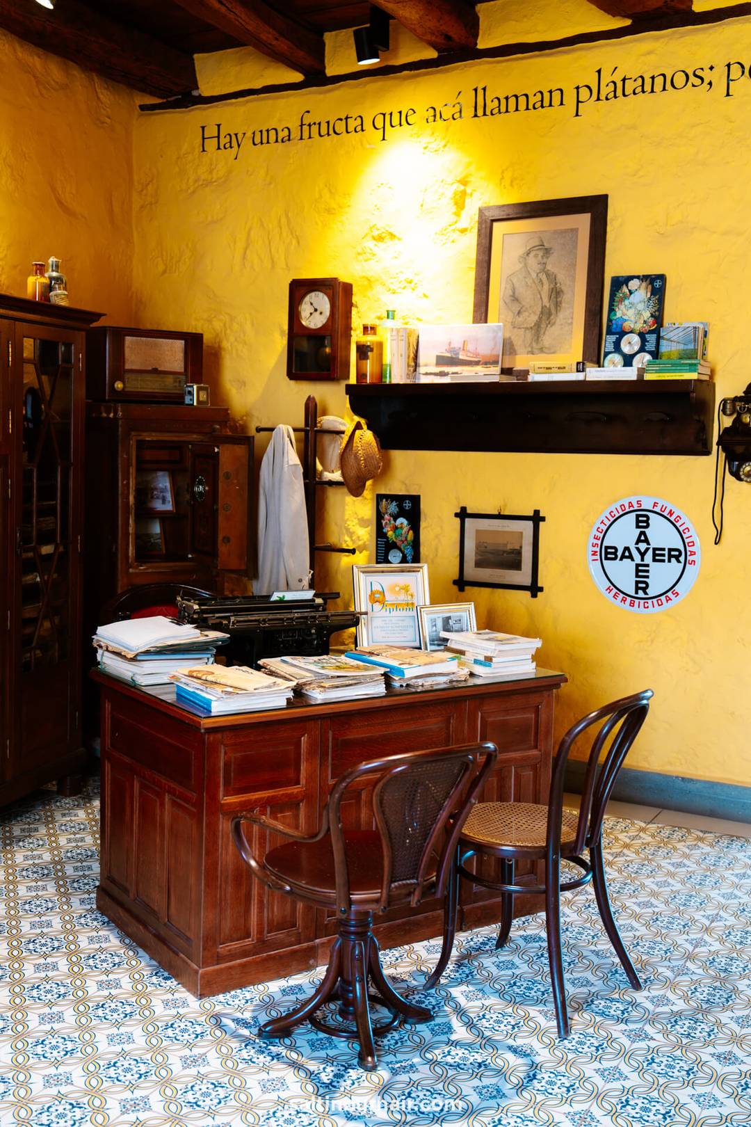 A vintage office with a wooden desk, two chairs, books, a typewriter, and framed photos sits against a yellow wall with shelves, clocks, and a Bayer sign. Spanish text is painted above in the Casa del Pl&aacute;tano (Banana Museum).