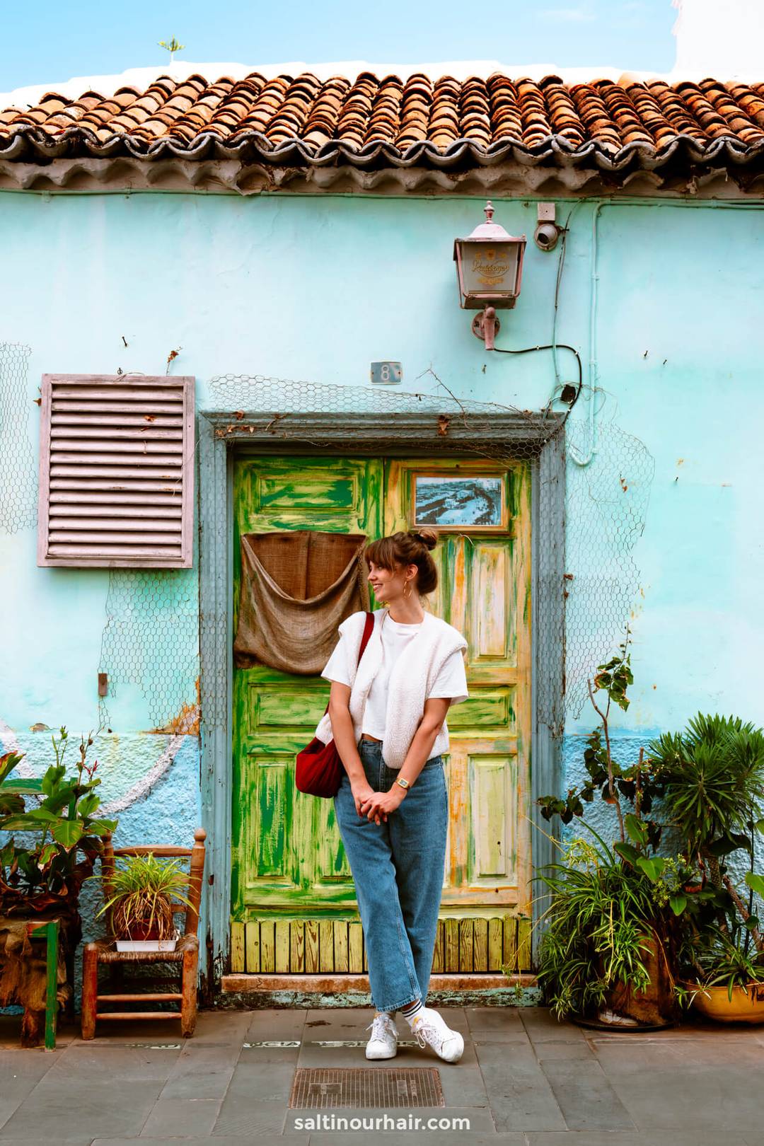 A woman stands in front of a rustic house with colorful green doors and potted plants in Puerto de la Cruz, Tenerife, wearing blue jeans, a white top, and white sneakers.