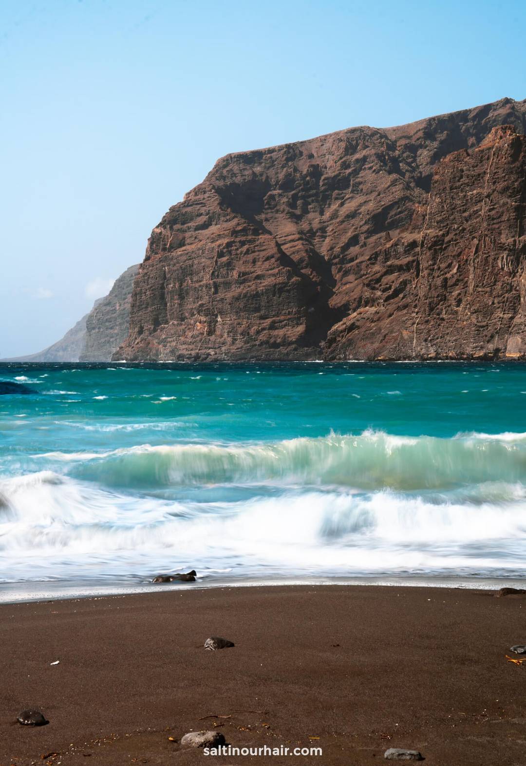 Waves wash onto the dark sandy shore of Los Gigantes beach Tenerife, with turquoise water and a large rocky cliff in the background under a clear sky.