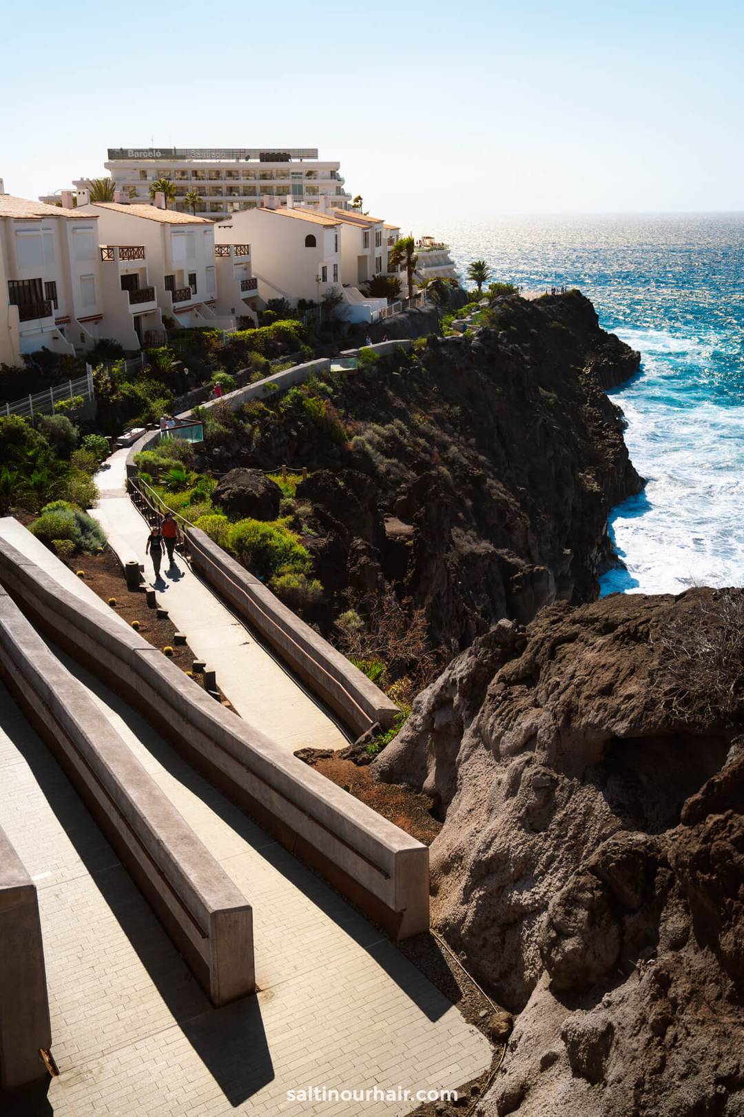 Two people stroll along a coastal cliff walk beside modern white buildings atop the rocky heights of Los Gigantes, Tenerife, overlooking the ocean on a sunny day.