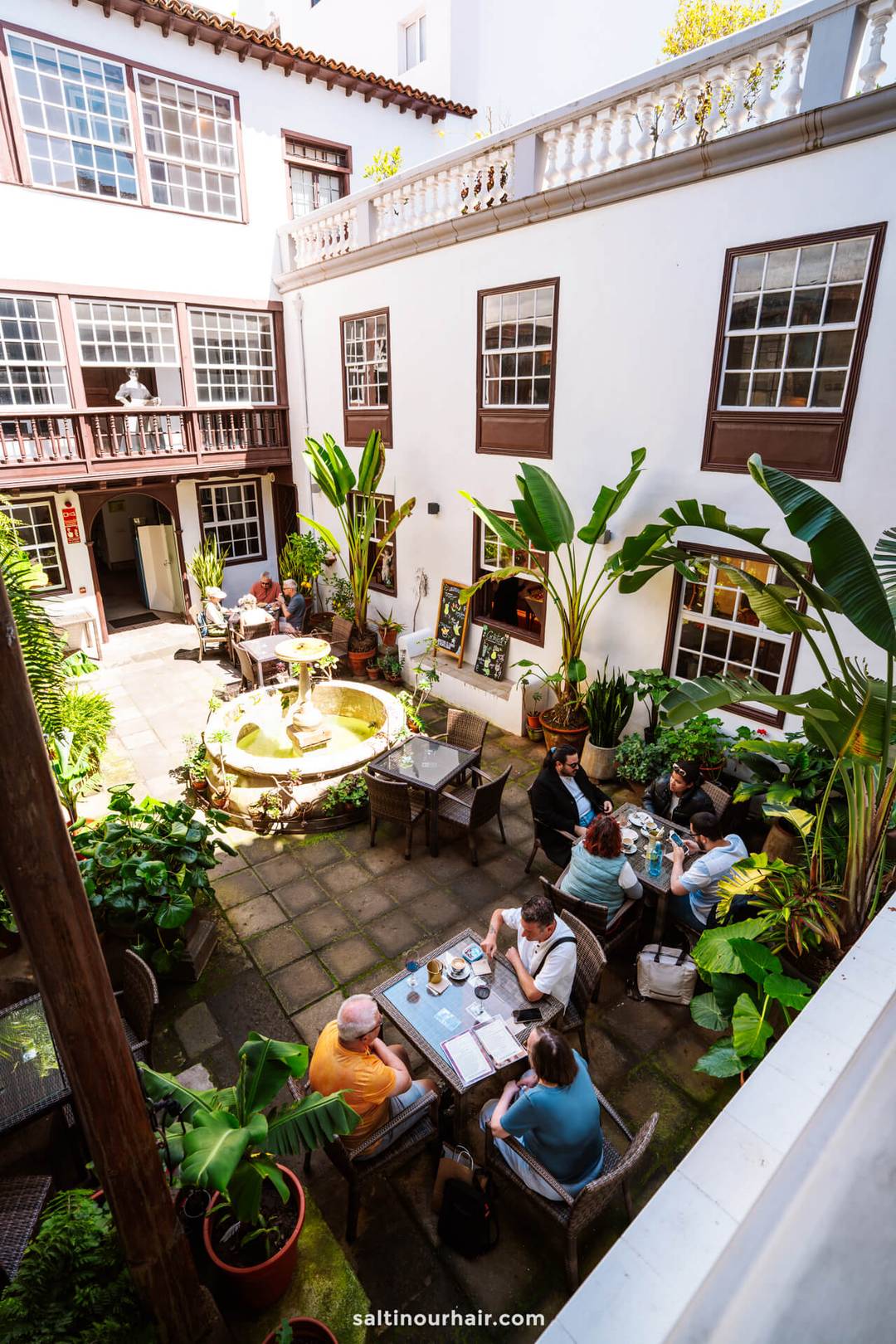 Een charmant terras op een binnenplaats in Casa Museo Cayetano G&oacute;mez in San Crist&oacute;bal de La Laguna, Tenerife, met mensen aan tafeltjes, omringd door weelderige planten en een centrale fontein.