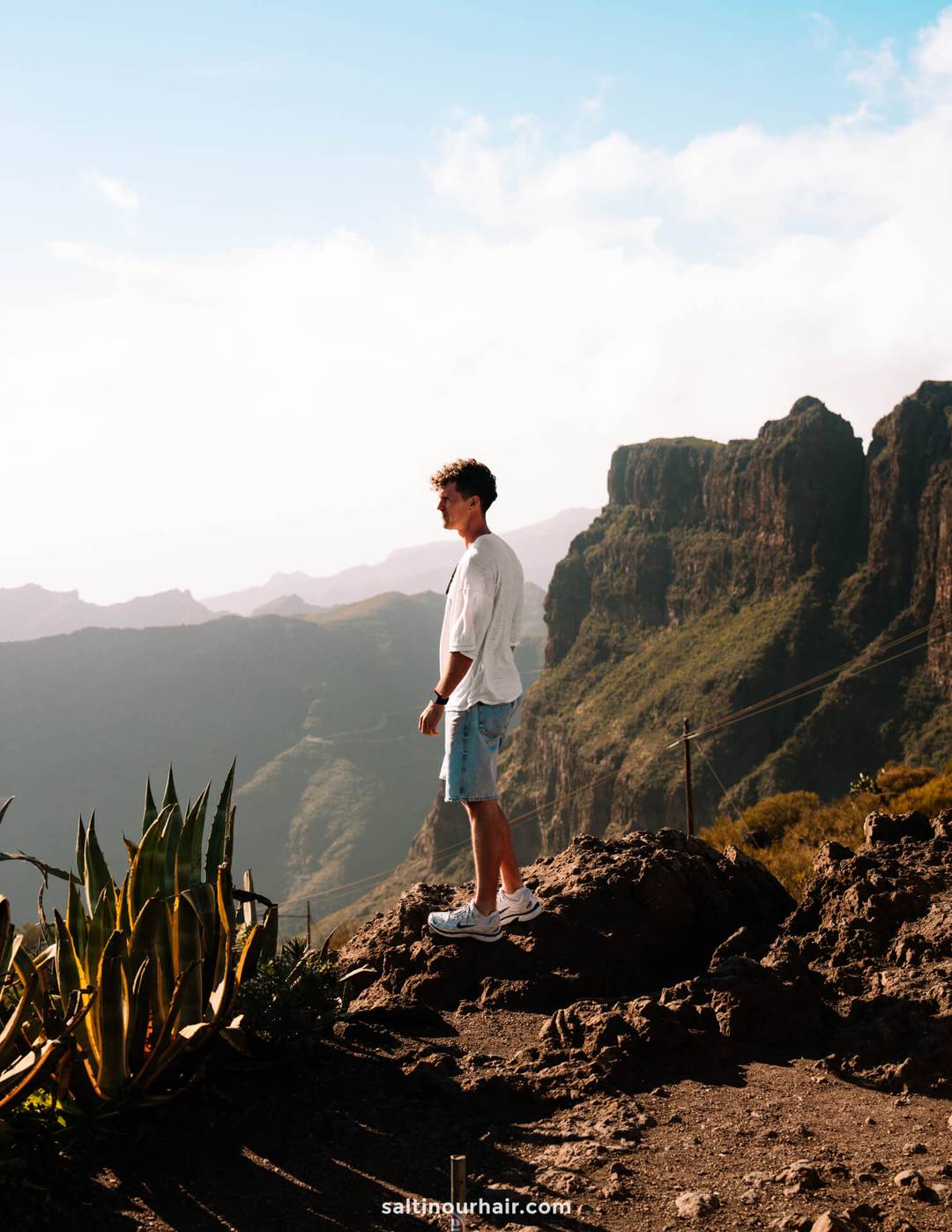 A man in a white shirt and shorts stands on a rocky outcrop, overlooking the stunning mountainous landscape of Masca, Tenerife, under a partly cloudy sky.
