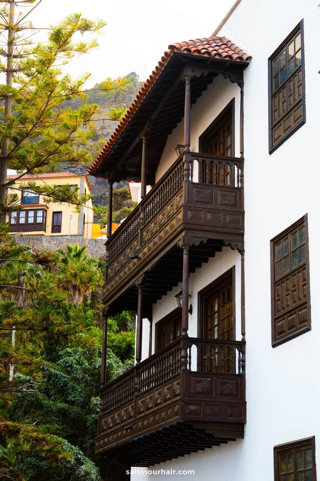 White building with two dark wooden balconies, detailed railings, and large windows, set against greenery and a hillside in the background&mdash;typical of Garachico Tenerife&rsquo;s charming architecture.