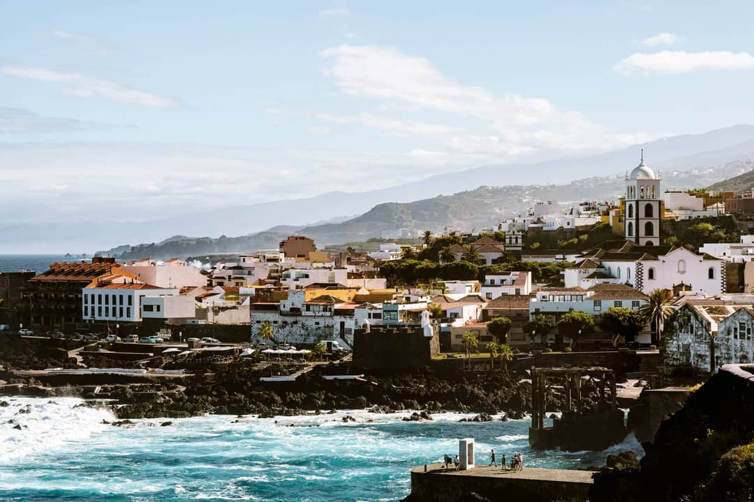 Coastal town of Garachico, Tenerife, with white buildings, a church with a bell tower, rocky shoreline, and waves; mountains and clouds in the background.