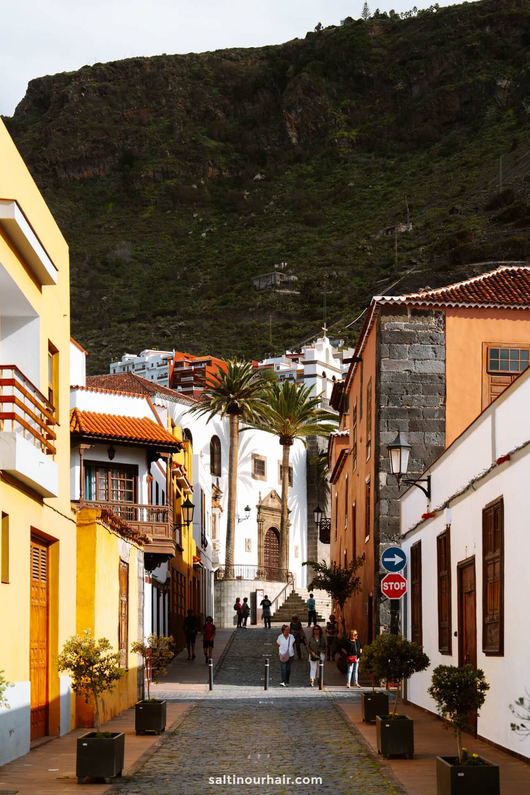 A cobblestone street with colorful buildings leads to a white church in Garachico, Tenerife, with people walking and a green mountain in the background.